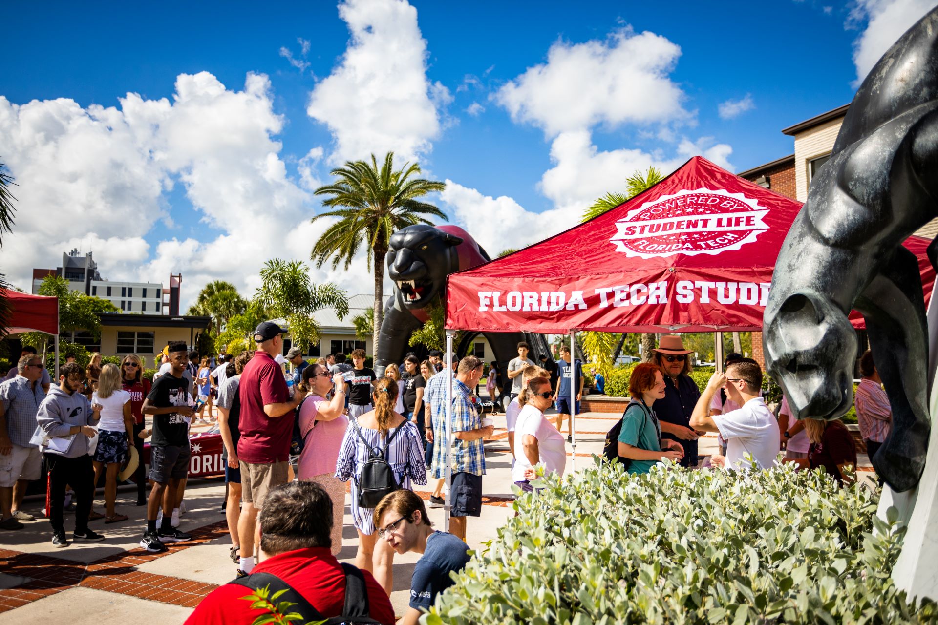 Students and visitors gather under a Florida Tech Student Life tent in Panther Plaza, with a large inflatable panther in the background, enjoying a sunny day with blue skies and palm trees